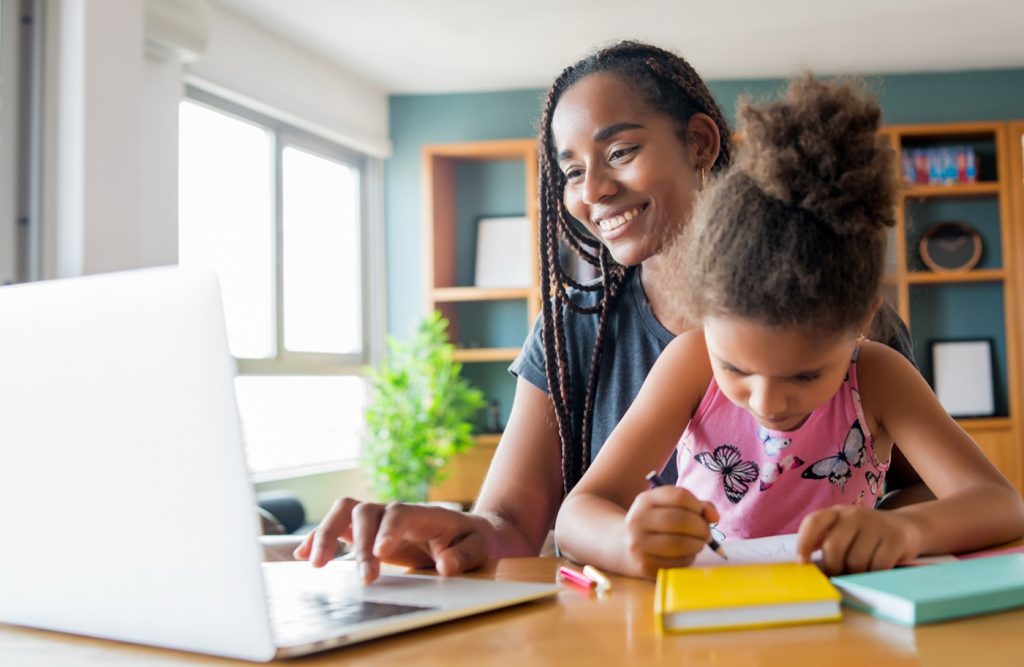 A mother leaning over her young daughter's shoulder at the laptop, guiding her as she writes in an exercise book.