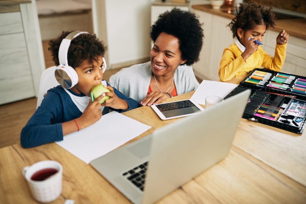 A mother laughing with her son as they work through a lesson on the laptop, while his sister draws beside them at the kitchen table.