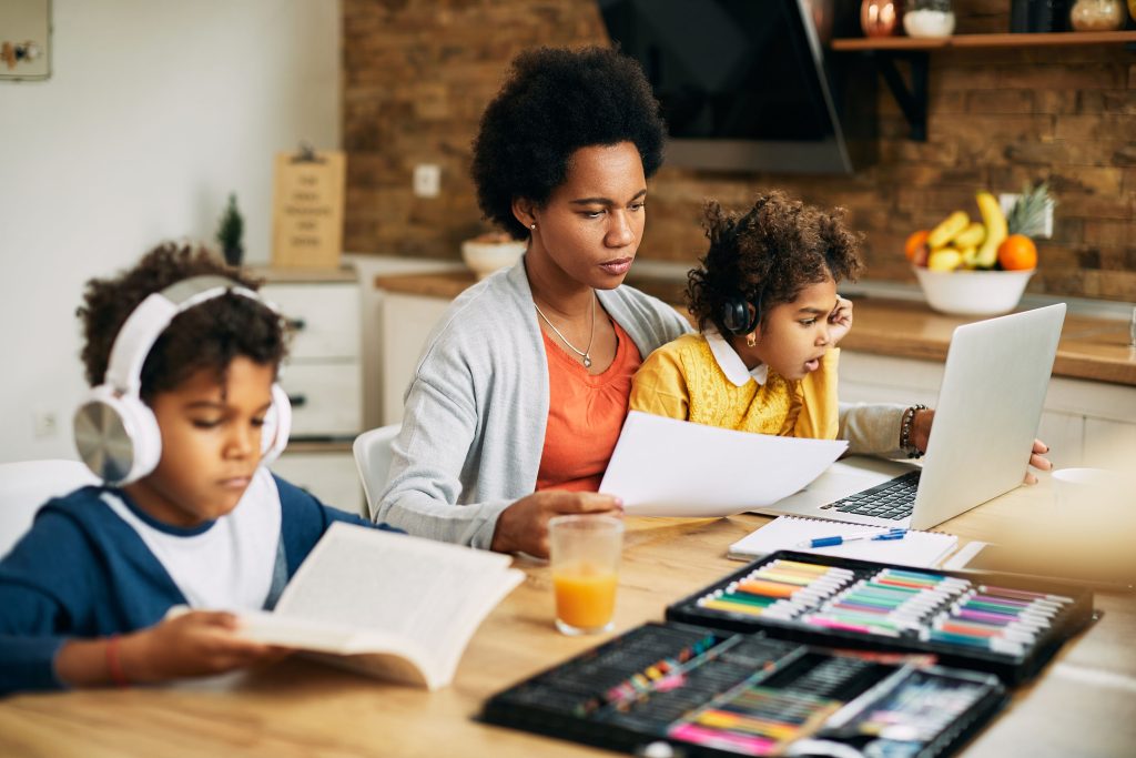A mother helping her daughter follow a lesson on the laptop while her son reads with headphones on at the same table.